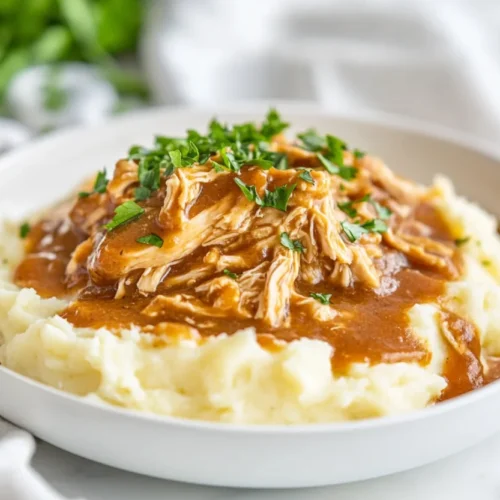 Slow cooker chicken and gravy in a white bowl garnished with parsley, served with mashed potatoes on a wooden board