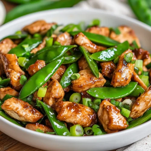 Chicken stir fry with snow peas on a white bowl, garnished with green onions and bright vegetables