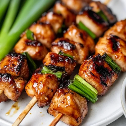 Grilled negima yakitori skewers on a white plate, glazed and garnished, with a bowl of sauce
